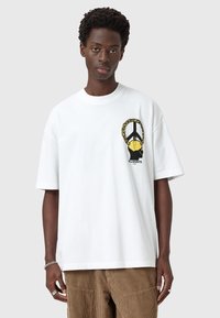 Hvid bomulds t-shirt med et sort og gult grafisk motiv på venstre bryst, med løs pasform og rund halsdesign.