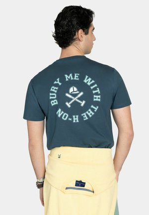 Hombre con cabello rizado oscuro que lleva una camiseta azul con bates de cricket cruzados y un casco, con el texto "Entiérrame con el H-ON" en la parte posterior.