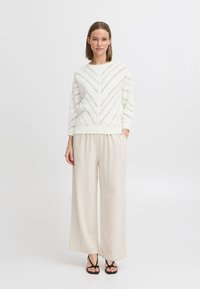 Weißer, gestrickter Pullover mit Zickzackmuster, kombiniert mit hellbeigen, weiten Hosen und schwarzen Sandalen mit Riemchen.