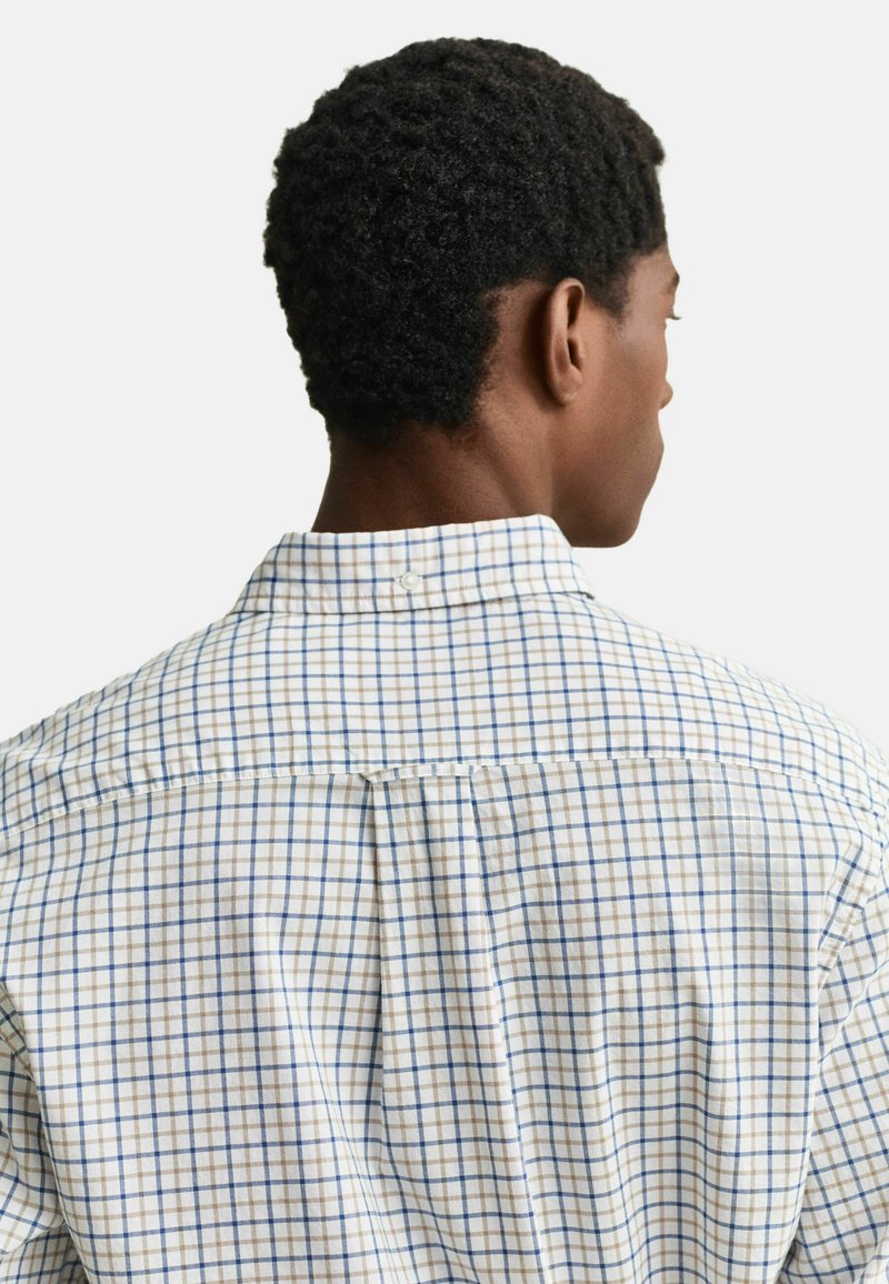 Homme aux cheveux courts et bouclés noirs, portant une chemise blanche à boutons avec un motif à carreaux bleu et marron, vu de dos.