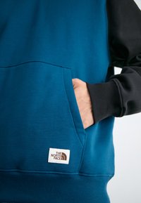 Felpa blu e nera con tasca a marsupio, polsini a coste e un piccolo etichetta di marca. Texture liscia, design casual.