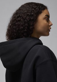 Sweatshirt preto com um capuz grande e versátil, feito de um tecido texturizado. Apresenta um corte descontraído e detalhes de costura visíveis ao longo dos ombros.