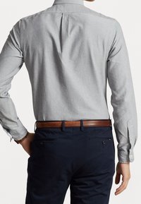 Uomo che indossa una camicia a maniche lunghe grigia chiaro infilata nei pantaloni blu navy con una cintura in pelle marrone, visto da dietro.