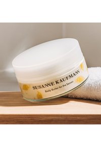 Beurre corporel dans un pot en verre transparent avec un couvercle blanc. L'étiquette présente des illustrations d'oiseaux jaunes et du texte noir. Serviette blanche douce à côté.