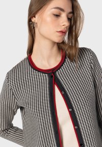 Cardigan a quadri in bianco e nero con bordi rossi e blu navy. Presenta una scollatura rotonda, chiusura frontale con bottoni e tessuto testurizzato.
