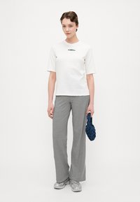 Hvid t-shirt med korte ærmer og et grønt logo, kombineret med sort-hvide stribede brede bukser og en struktureret marineblå clutch. Sølv sneakers.