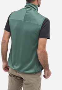 Grüne, ärmellose Weste mit hohem Kragen. Texturiertem Stoff mit einem dezenten Muster. Getragen über einem schwarzen T-Shirt mit kurzen Ärmeln und beigen Hosen.