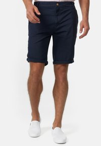 Shorts en coton bleu marine avec des ourlets retroussés, des poches avant et une fermeture à bouton. Portés avec des chaussures blanches à enfiler sur un fond neutre.