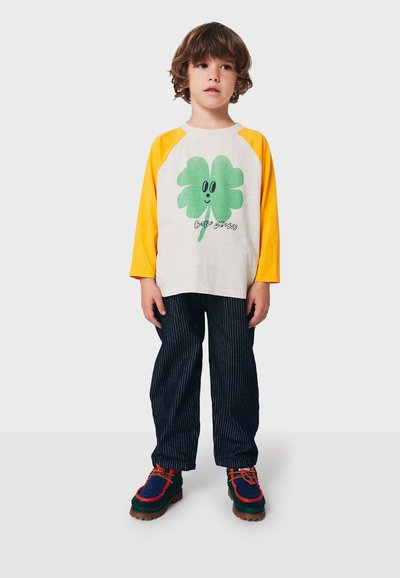 Long-sleeve raglan shirt with grey body and yellow sleeves, featuring a green clover graphic. Striped dark blue pants and colorful shoes.