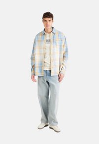 Camicia a quadri blu chiaro e gialla sopra una maglietta beige, abbinata a jeans blu chiaro e sneakers bianche. Design casual e dalla vestibilità rilassata.