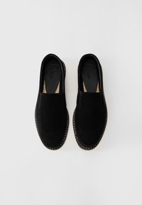 Une paire de chaussures slip-on en daim noir avec des coutures autour de la semelle, présentée sur un fond clair et uni.