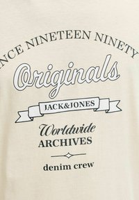 Beige katoenen T-shirt met ronde hals, met zwarte grafische tekst: "Sinds Negentien Negentig," "Originals," en "Jack & Jones" met decoratieve bannerdetails.