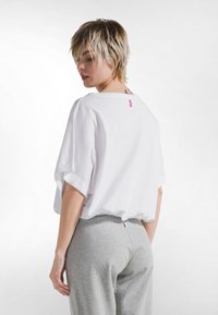 Camiseta blanca de algodón extragrande con mangas cortas remangadas, etiqueta rosa en la parte posterior, combinada con pantalones de chándal grises, textura suave, diseño casual.