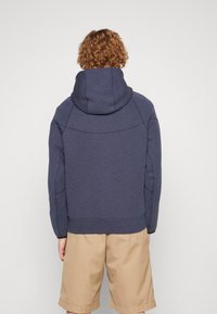 Felpa blu navy con colletto alto e cappuccio ampio. Realizzata in tessuto testurizzato, presenta maniche raglan e polsini elasticizzati. Indossata con pantaloni corti beige.