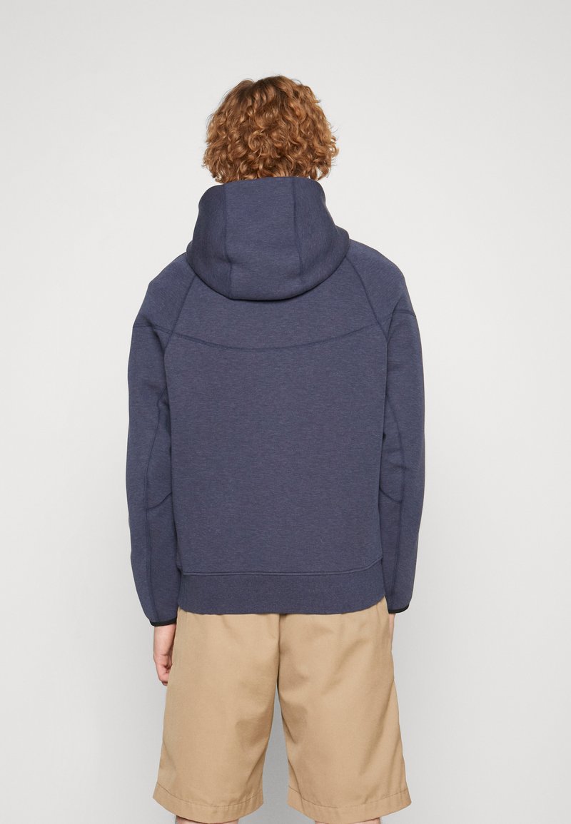 Sweat à capuche bleu marine avec un col haut et une grande capuche. Fabriqué en tissu texturé, avec des manches raglan et des poignets élastiques. Porté avec un short beige.