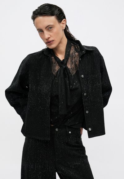 Giacca di jeans nera con texture scintillante e chiusura a bottoni, sovrapposta a una blusa in pizzo nero trasparente con collo annodato, abbinata a pantaloni coordinati.