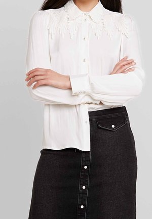 Femme portant un chemisier blanc à manches longues avec col en dentelle et boutons, associé à une jupe en jean noire taille haute avec boutons sur le devant.