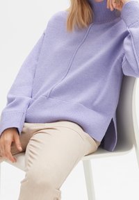Personne assise sur une chaise blanche portant un pull en tricot lavande à col roulé et un pantalon beige, reposant un bras sur le dossier de la chaise.