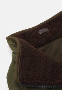 Jaqueta verde-oliva com gola forrada em fleece castanho. Detalhe de fecho zip na lateral. Apresenta etiqueta da marca no tecido interior.