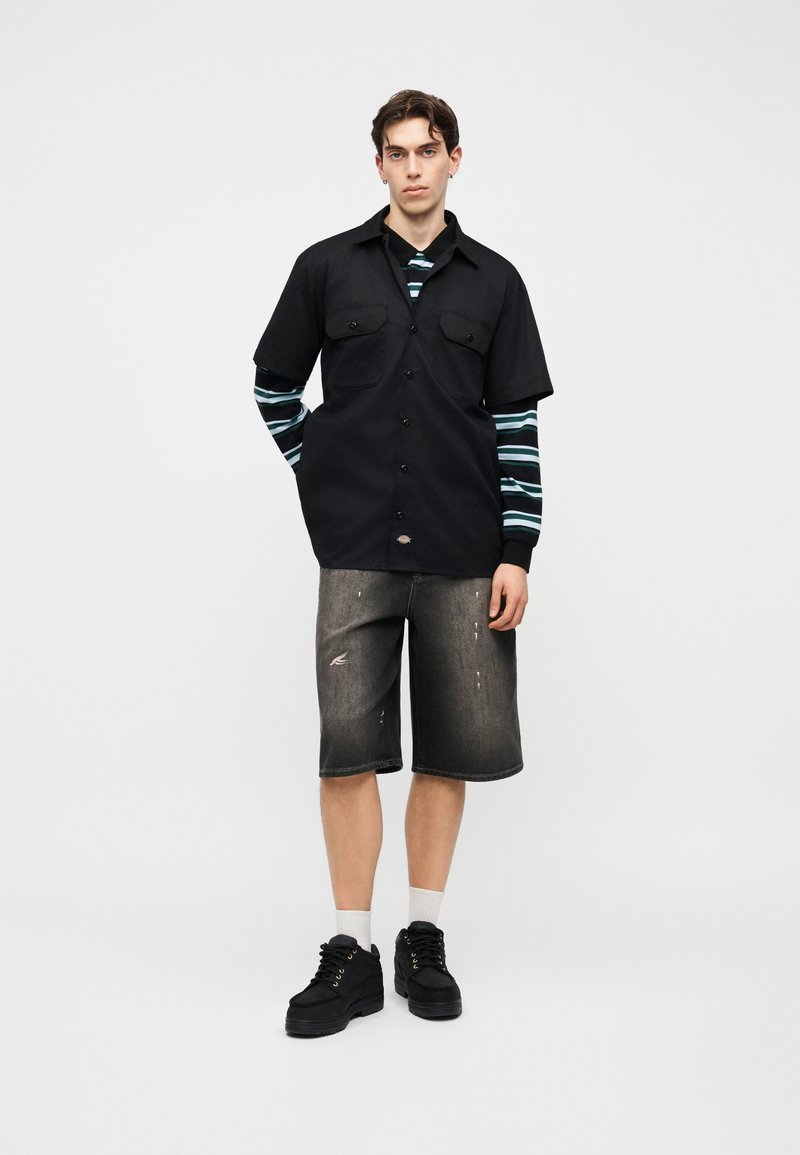 Junger Mann trägt ein schwarzes kurzärmeliges Hemd mit Knöpfen über einem gestreiften langärmeligen Shirt, ausgewaschene schwarze knielange Shorts, weiße Socken und schwarze Schuhe.