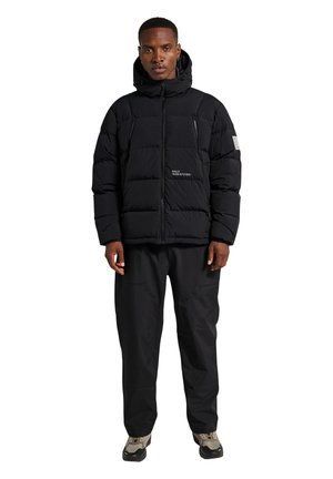 Schwarze gepolsterte Jacke mit einem Reißverschluss vorne, ausgestattet mit einer Kapuze, Seitentaschen und Logodetails. Getragen mit schwarzen Hosen und gemustertem Schuhwerk.