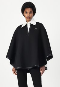 Juodas pončas su balta apykakle, turintis ketvirčio užtrauktuką, su lygaus paviršiaus audiniu ir kontrastinėmis odos primenančiomis detalėmis.