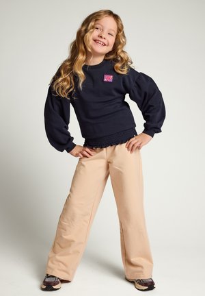 Jong meisje met lang blond haar poseert glimlachend, draag een marineblauwe sweatshirt met een roze patch, beige wijde broek en sneakers.
