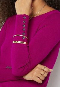 Fuksjowy sweter z długimi rękawami, ozdobiony sześcioma czarnymi guzikami przy mankiecie; na nadgarstku noszone złote i srebrne bransoletki.