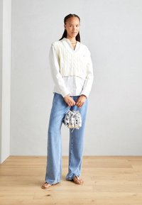 Chaleco corto de punto blanco sobre una camisa blanca abotonada, combinado con pantalones vaqueros de pierna ancha azul claro y una bolsa metálica de cordón.
