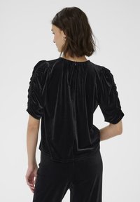 Top in velluto nero con maniche corte a sbuffo, chiusura a occhiello sul retro e dettagli arricciati sulle spalle. Tessuto morbido con una leggera lucentezza.