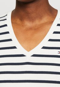 Bílý tričko s v-výstřihem a námořnickými horizontálními pruhy, s krátkými rukávy a texturovanou pleteninou. Malé logo je na levé straně.
