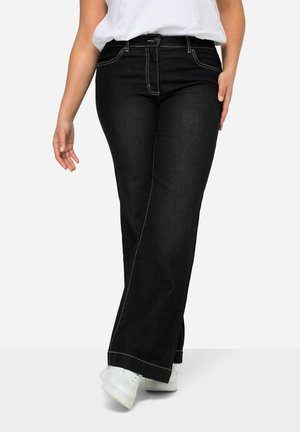 Jeans en denim noir évasés avec des coutures contrastantes, cinq poches, fermeture à bouton et à zipper, et une texture lisse. Portés avec des chaussures blanches.