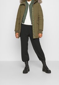 Olivgrön parka med fuskpälshuva, kviltad innerskikt, svarta vida byxor och kraftiga svarta snörkängor. Texturerade material synliga.