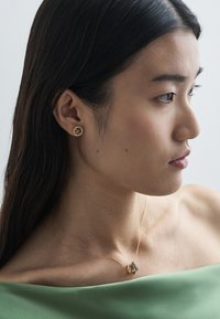 Zlaté náušnice s kulatým designem a barevnými akcenty; jemný zlatý náhrdelník s malým geometrickým přívěskem. Modelka má na sobě zelený top.