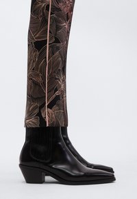 Stivaletti alla caviglia in pelle nera con punta affilata e tacco a blocco, abbinati a pantaloni con motivo floreale in rosa chiaro su sfondo nero.