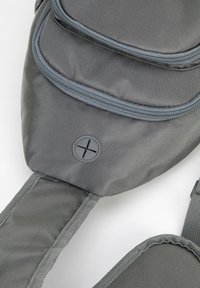 Szara torba na ramię z tkaniny o fakturowanej powierzchni, dwoma kieszeniami zapinanymi na zamek oraz okrągłym logo. Regulowany pasek zapewniający wygodne noszenie.