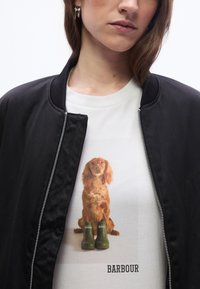 Γυναίκα που φορά μαύρο μπουφάν πάνω από λευκή μπλούζα με εικόνα καφέ σκύλου που κάθεται πίσω από δύο πράσινες μπότες Barbour με το λογότυπο Barbour από κάτω.