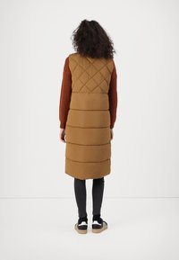Gilet long rembourré marron avec un quiltage en losanges sur les épaules et un ourlet droit. Porté sur un pull couleur rouille et un jean sombre, associé à des baskets noires.
