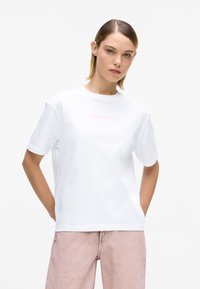 Valge puuvillane T-särk ümmarguse kaelusega, lühikeste varrukatega ja keskel roosade logotekstidega. Kandmisel heleroosad teksapüksid.