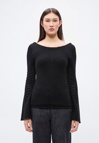 Fekete kötött felső széles nyakkivágással és hosszú, bővülő ujjakkal. A testén texturált halszálkaminta látható. Sötét nadrággal párosítva.