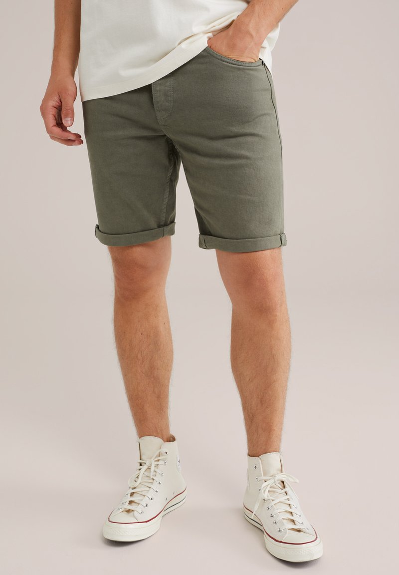 Mies, joka on pukeutunut ylös käännettyihin oliivinvihreisiin shortseihin, valkoisiin korkeisiin tennareihin ja valkoiseen paitaan, seisoo tasaisella taustalla toinen käsi taskussa.