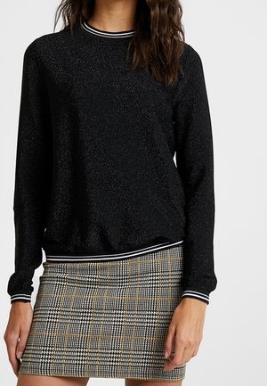 Femme portant un pull noir à manches longues avec des poignets et un col rayés blancs, associé à une mini-jupe à carreaux à motifs.