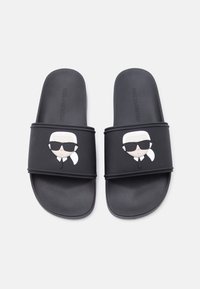 Sandales à glissière noires avec des visages de dessin animé portant des lunettes de soleil et des queues de cheval blanches sur les lanières, semelles texturées et nom de la marque embossé.