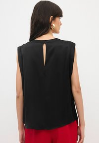 Blusa nera senza maniche con scollatura tonda, apertura a forma di occhio di bue sul retro e una texture liscia, abbinata a pantaloni rossi.