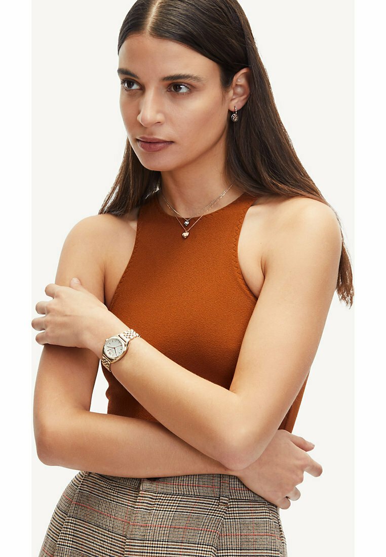 Ackerrötiger ärmelloser Top mit strukturierter Oberfläche, kombiniert mit einer silberfarbenen Armbanduhr und geschichteten Halsketten. Die Hand des Models ruht auf ihrem Arm.