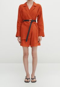 Femme portant un blazer orange à manches longues ceinturé et un short assorti avec des sandales plates noires sur fond blanc.