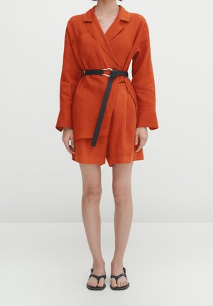 Femme portant un blazer orange à manches longues ceinturé et un short assorti avec des sandales plates noires sur fond blanc.