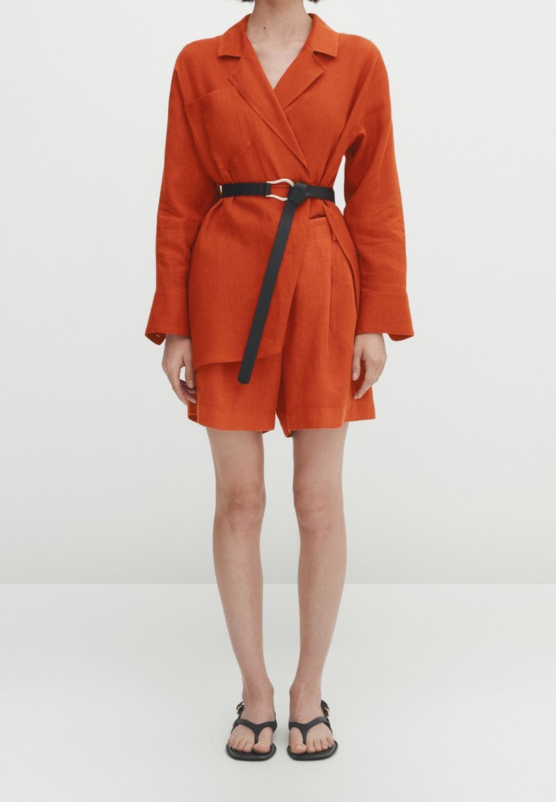 Femme portant un blazer orange à manches longues ceinturé et un short assorti avec des sandales plates noires sur fond blanc.