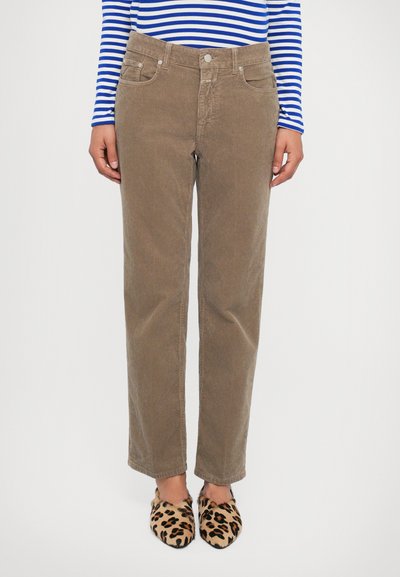 Persona vestida con pantalones rectos de pana beige, una camiseta de rayas azul y blanca de mangas largas, y zapatos tipo slip-on con estampado de leopardo, de pie contra un fondo blanco.