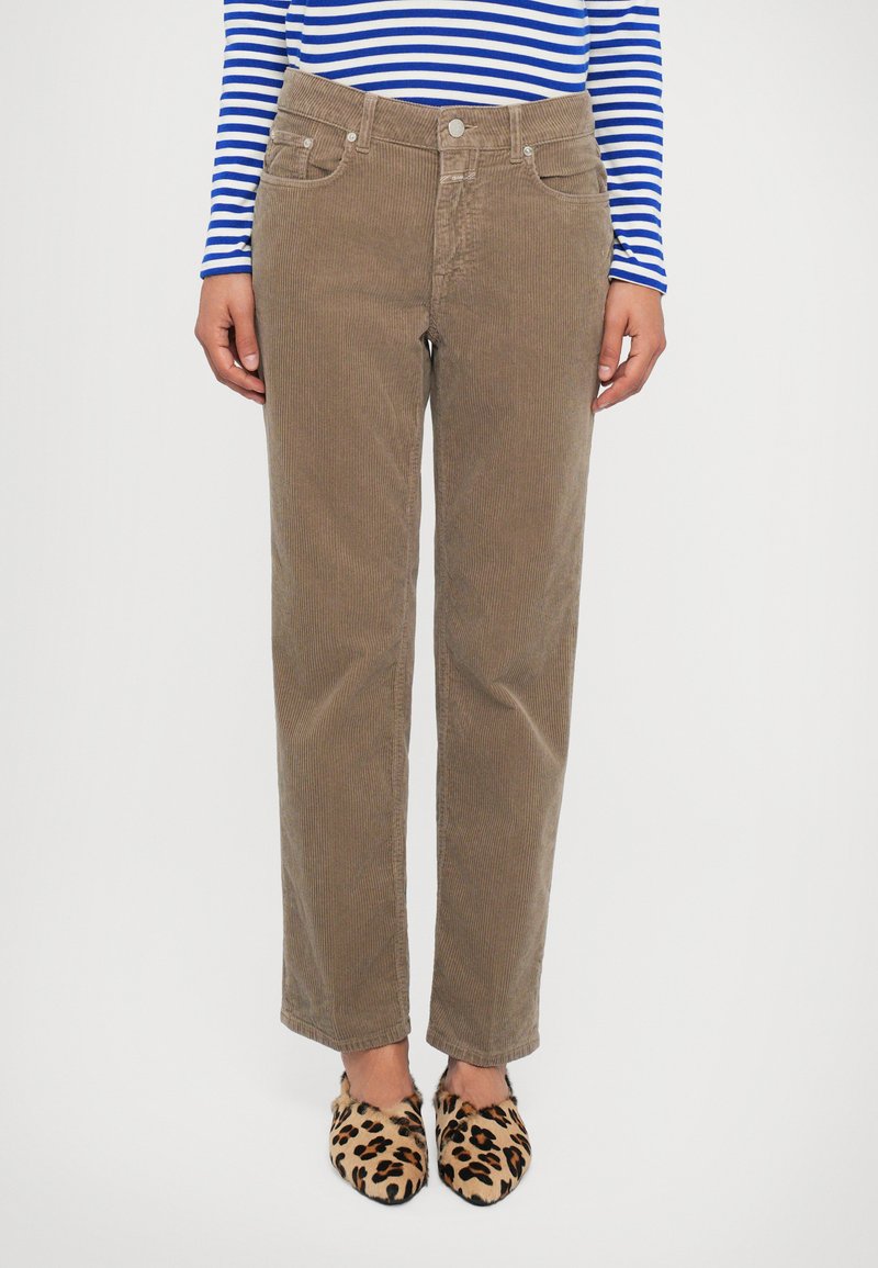 Persoon in beige corduroy straight-leg broek, met een blauw en wit gestreept langarmtop en luipaardprint instapschoenen, staand tegen een witte achtergrond.